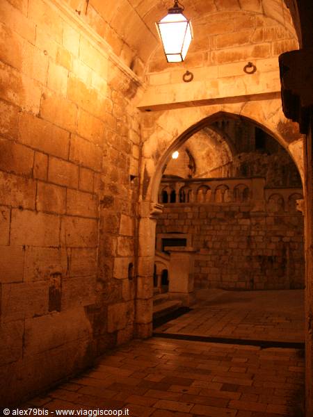 Lit lantern under a stone archway, glimpse of a medieval loggia beyond
