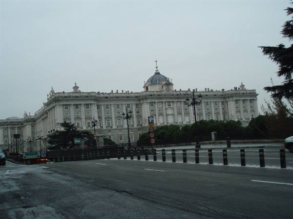 Royal Palace of Madrid under grey sky, wet road and bollards in front