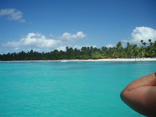 Arm in foreground over turquoise water facing Saona beach lined with palms