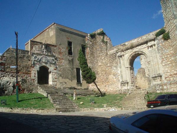 Stone ruins of old convent with portal arch, staircase, blue sky, parked cars