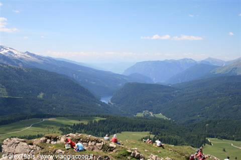 Hikers resting on grassy rocks overlook a valley with a blue lake