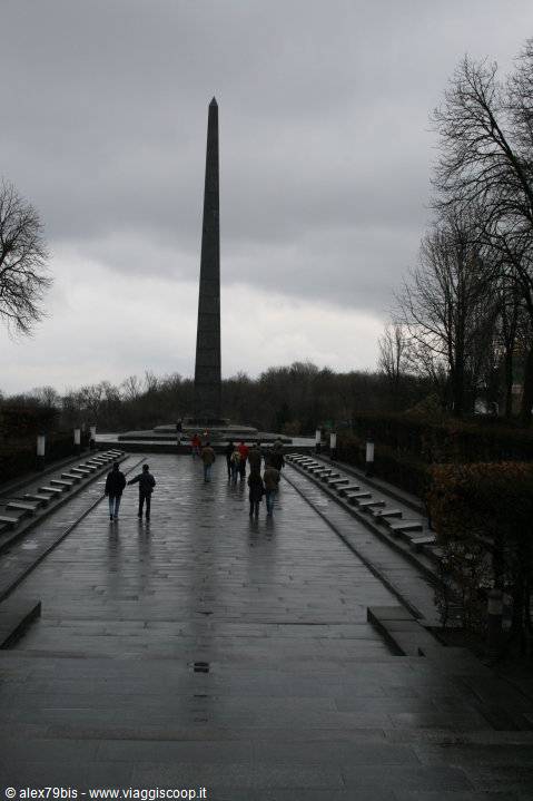 Visitors walk along the wet promenade toward the memorial obelisk, gray sky