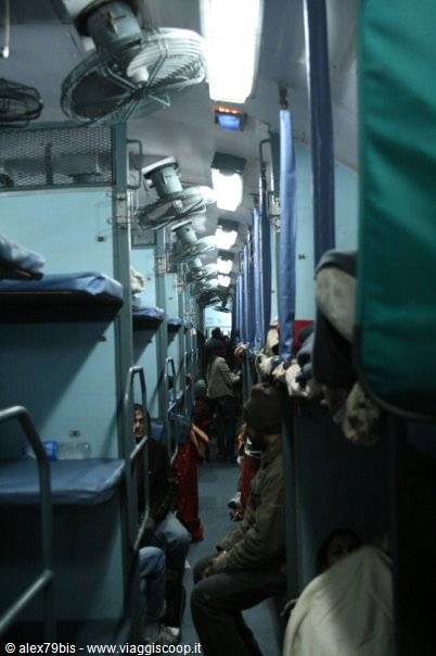 Crowded sleeper train carriage with ceiling fans and neon lights, passengers on bunks