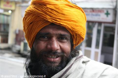 Man with orange turban and beard smiling on a street among shops