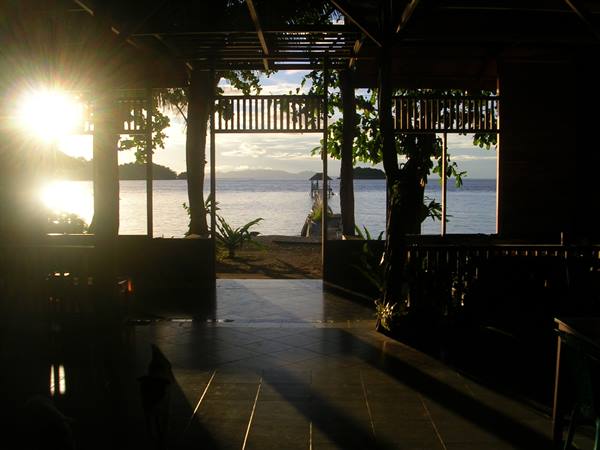 Low sun streams through wooden pavilion opening onto sea with pier