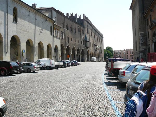 Piazza Sordello in Mantua, brick arcaded buildings, cars parked on cobblestones, daylight