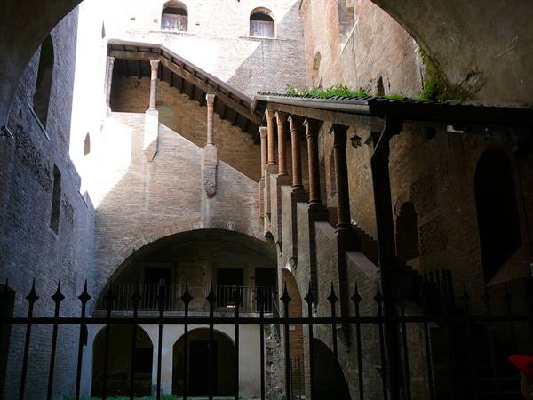 Brick courtyard with covered staircase and columned loggia behind a wrought-iron gate