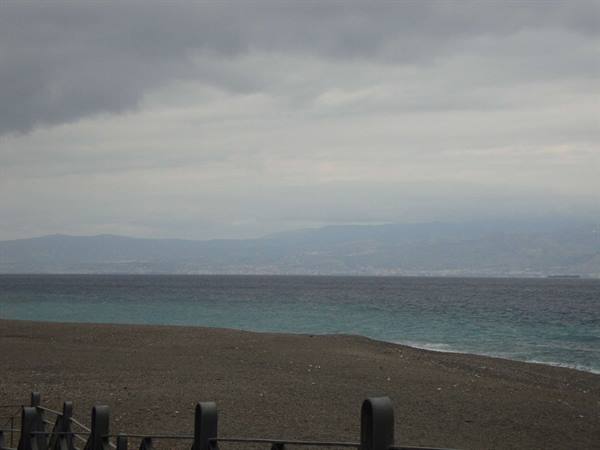 Pebble beach with metal railing, turquoise sea and mountains under cloudy sky