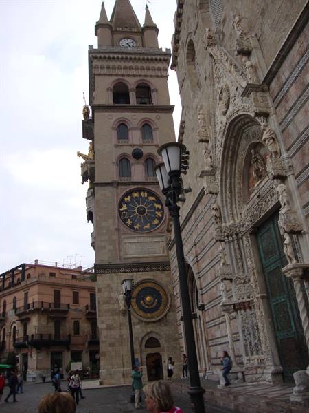 Bell tower with astronomical clock beside Messina Cathedral's carved facade, tourists walking by