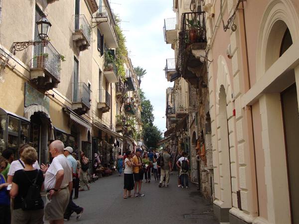 Tourists stroll past old buildings with flowered balconies and shops, Taormina