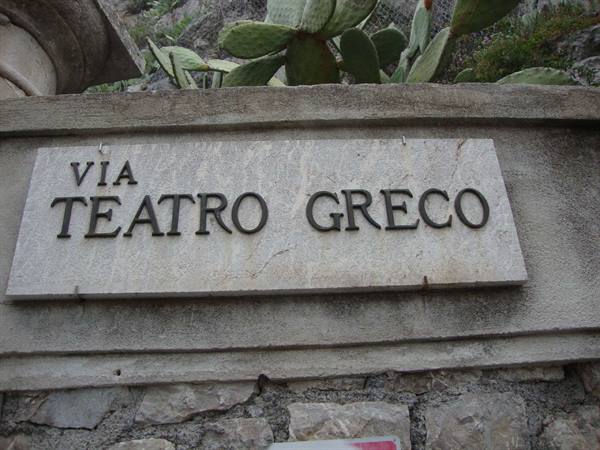 Marble plaque reading Via Teatro Greco on stone wall, prickly pears above