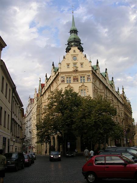 Baroque building with green spire in Prague, parked cars on cobblestones under clouds