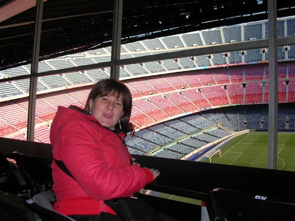 Woman in red jacket sits in Camp Nou stand, empty seats and green pitch