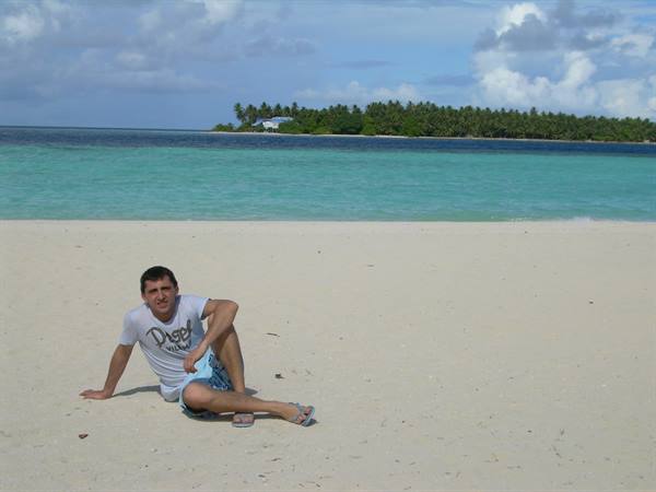 Guy sitting on white sand, turquoise lagoon and palm tree island