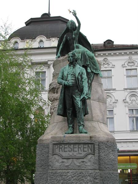 Green bronze statue of poet Prešeren with muse, in front of white Ljubljana palace