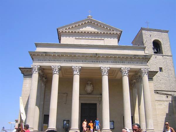Tourists on the steps of the Basilica of San Marino, Corinthian columns and bell tower under blue sky