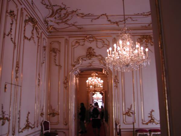 Lit crystal chandelier in gilded stucco hall with visitors at doorway