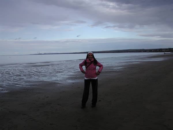 Woman in pink jacket on dark sandy beach at low tide under grey clouds