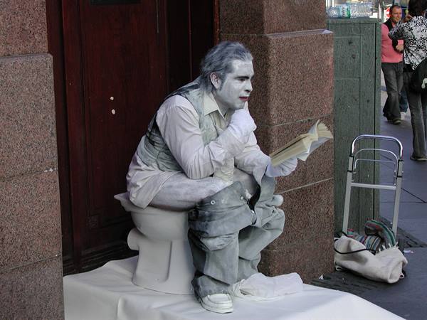 White-painted mime sitting on a toilet reading a book on the street