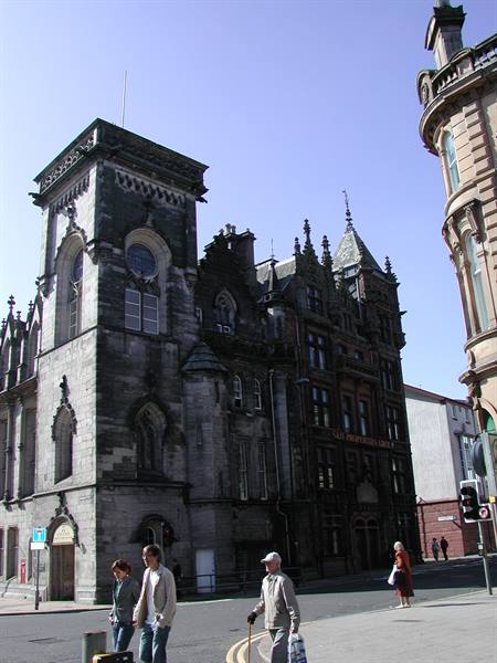 Pedestrians cross the street by a dark Victorian stone building