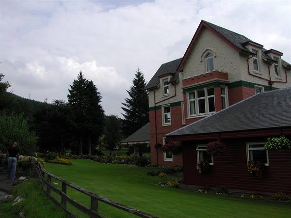 Person walking beside red-brick and white villa, green lawn and conifers, cloudy sky