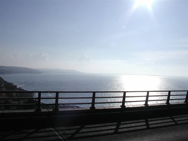 High sun over the sea, long causeway in the distance, dark railing in the foreground
