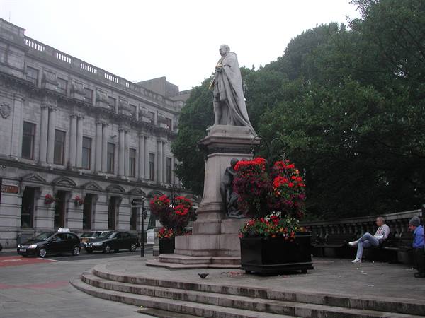 Stone statue on pedestal with red flower planters, classical building and passing taxis