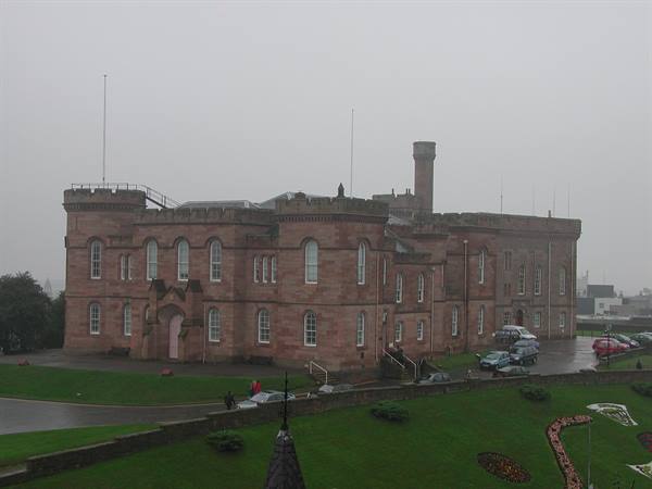 Red stone castle under grey rain, green lawn with flowerbeds and cars