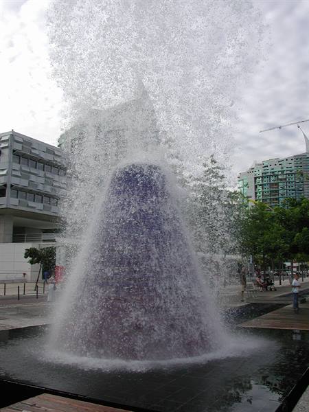 Cone-shaped fountain splashing in an urban plaza among modern buildings and trees