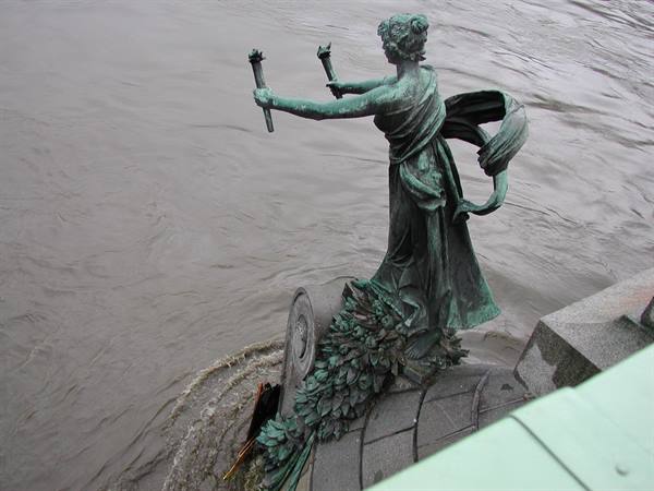 Green bronze statue holding two torches above the river in Prague