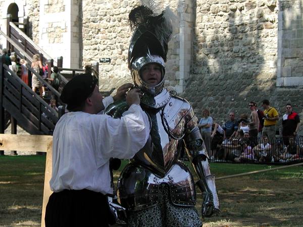 A man helps a knight in steel armor before a stone wall as tourists watch