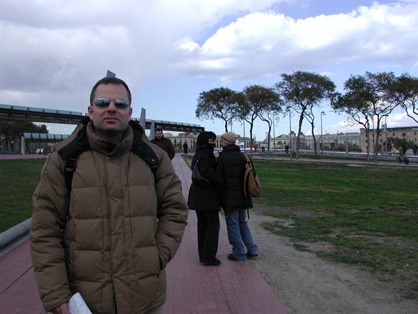Man in down jacket with backpack on paved walkway, two people behind among trees and grass