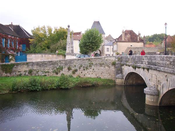 Tourists cross a stone bridge over the river, houses and obelisk behind