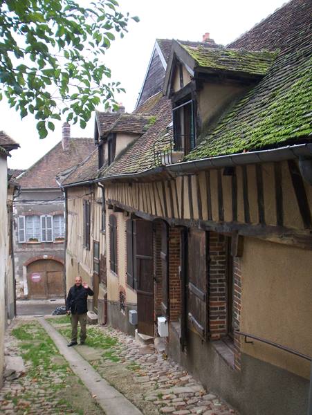 man waving beside half-timbered houses with mossy tile roofs and cobblestones
