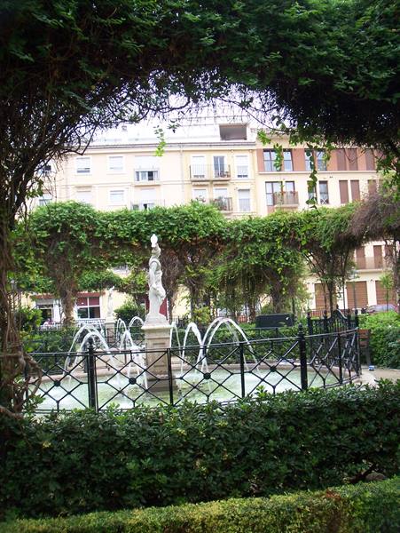 Fountain with statue in a city garden, beneath a green pergola among buildings