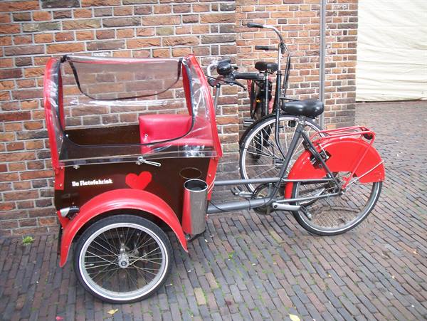Red De Flaterfietsch cargo trike parked next to a bicycle on brick pavement