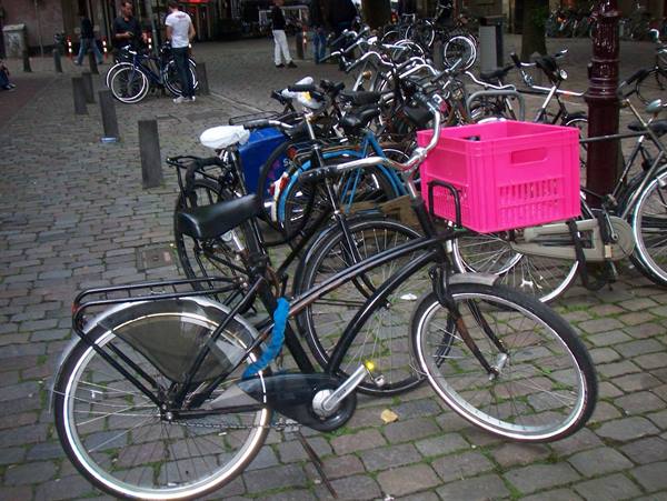 Black bicycles parked on cobblestones, one with pink crate, people nearby