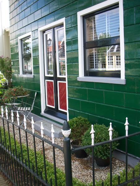 Glossy green wooden house with red door, iron fence, potted shrubs