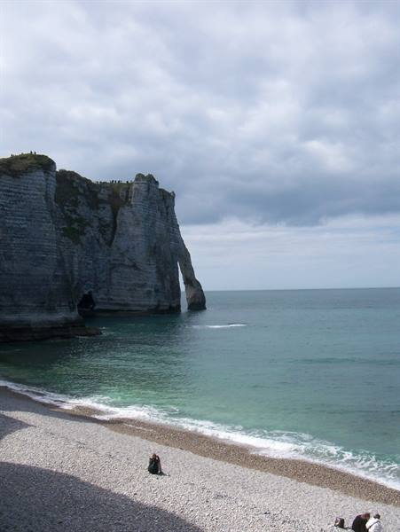 White chalk cliff with natural arch at Étretat, pebble beach, green sea under cloudy sky