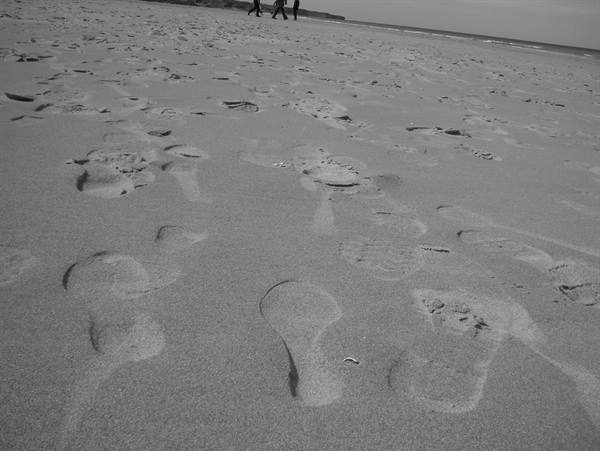 Footprints in grey sand, three distant walkers heading along the shoreline