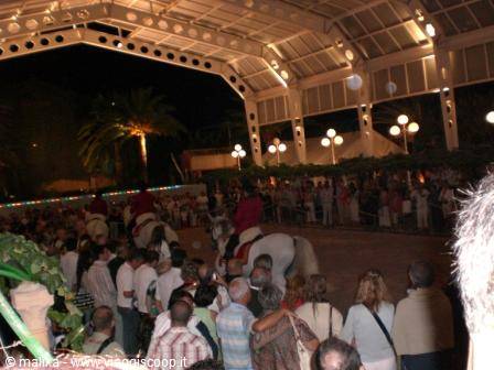 Riders on white horses among the crowd at a night fair under lit pavilion