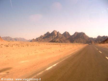 Paved road through sandy desert with rocky mountains in the background