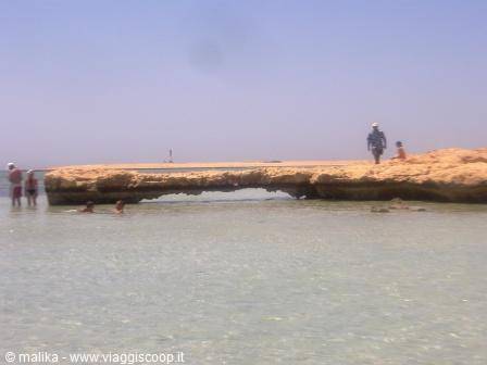 Tourists walk across a sandy rock ledge over turquoise water under bright sun