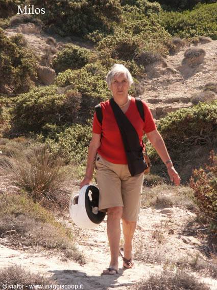 Woman with bag and white helmet walking on sandy path among bushes in Milos