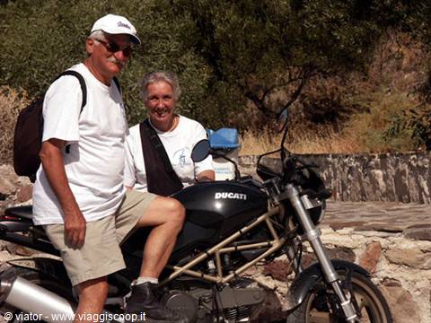 Couple posing beside a parked Ducati motorcycle on a dirt road among Mediterranean scrub