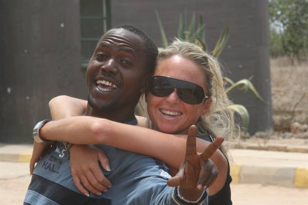 A man flashes a peace sign as a blonde woman in sunglasses hugs him smiling