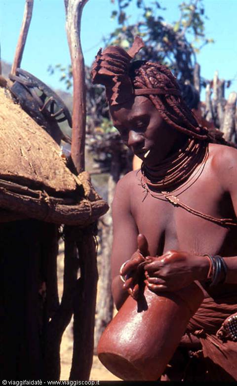 Himba woman with shell necklaces prepares food in a gourd by her hut