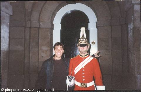 Smiling tourist next to a royal guard in red uniform under a stone arch