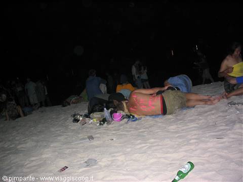Guy asleep on the sand at night, back painted pink, bottles scattered around