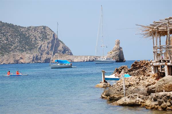 Sailboats anchored off Benirràs rock, canoes and a wooden hut on the rocky shore